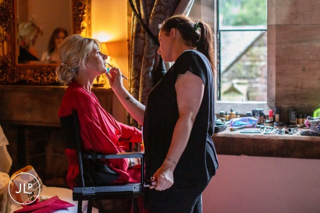Cheshire wedding photographer captures a makeup artist applying lipstick to the bride by a castle window.