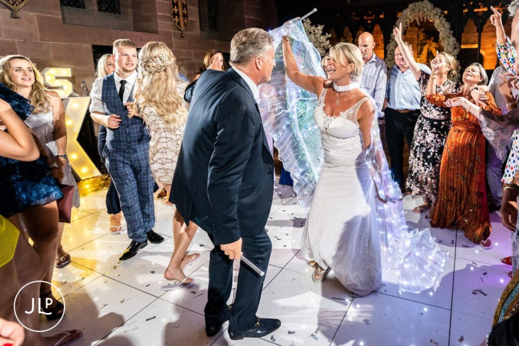 A bride in glowing LED wings dancing with her groom on a packed white dance floor in Peckforton Castle.
