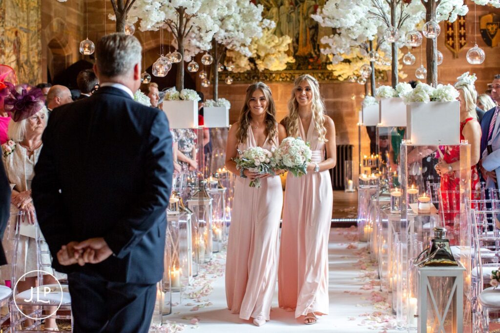Bridesmaids walking down the white aisle beneath floral trees at a beautiful Cheshire castle wedding.