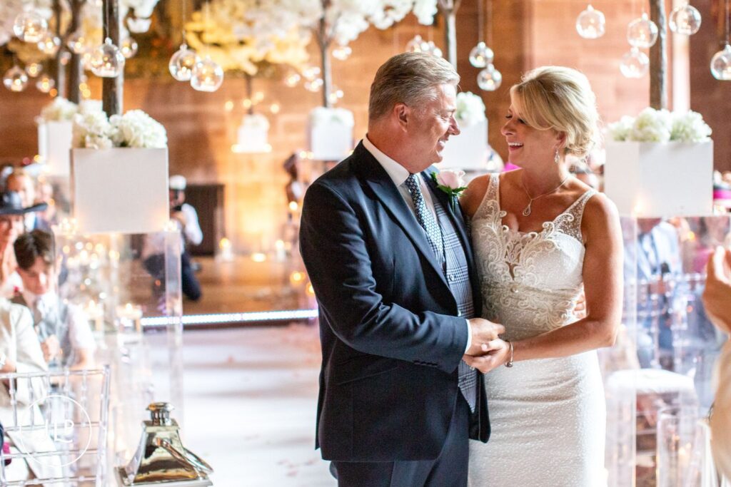 A bride and groom holding hands and smiling during their emotional Peckforton Castle wedding vows.