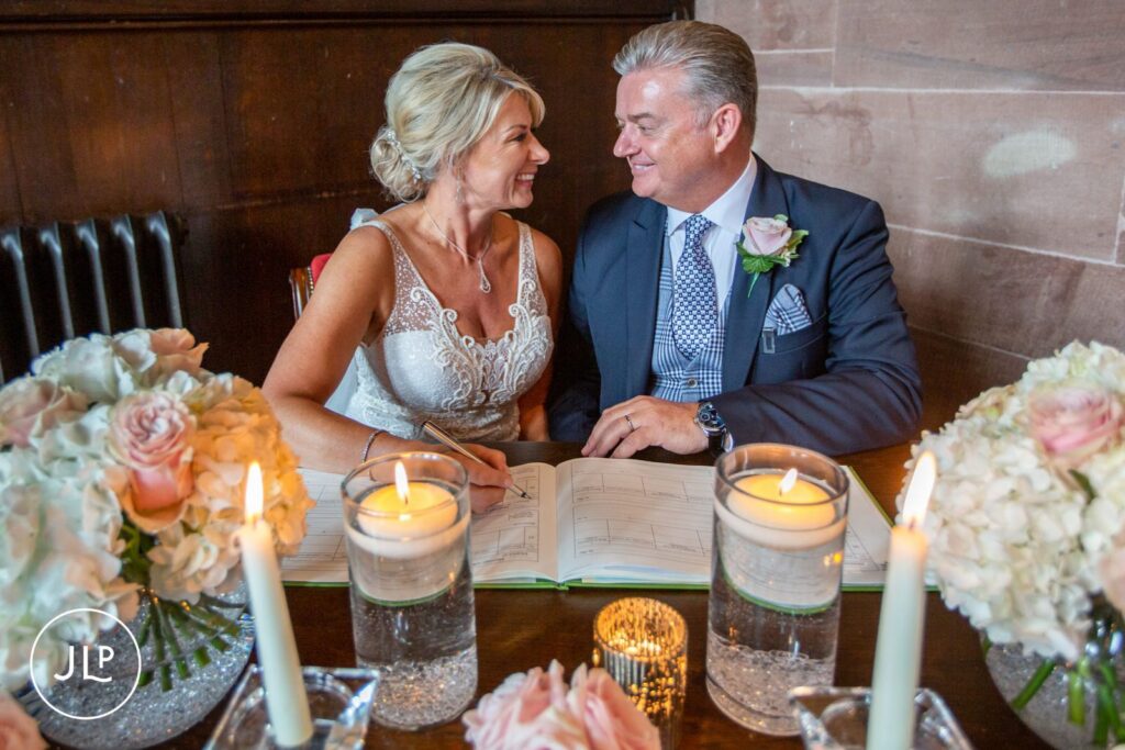 A bride and groom smiling at each other while signing the marriage register at a luxury castle wedding.