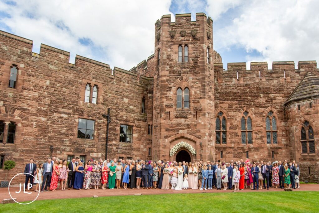 A wide group photo of guests outside the majestic historic architecture for a Peckforton Castle wedding.