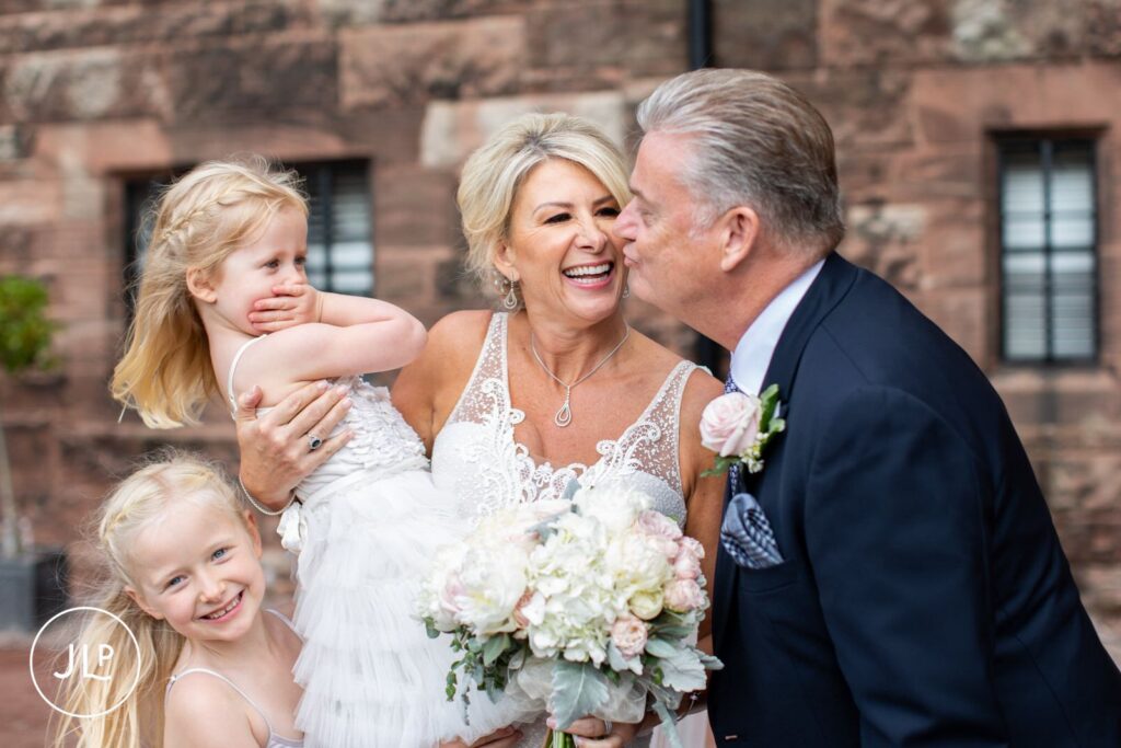 A groom playfully kisses his laughing bride while she holds two young flower girls in Cheshire.