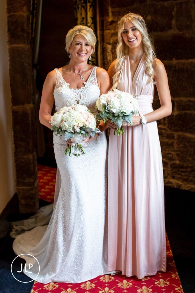 A smiling bride and bridesmaid holding white bouquets at a Peckforton Castle Cheshire wedding.
