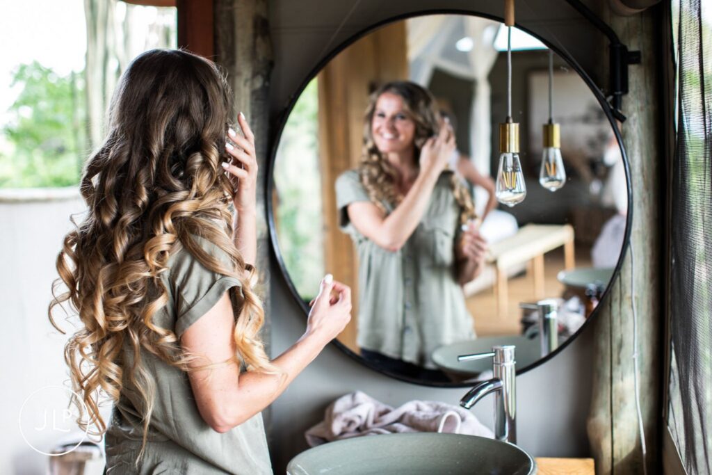 A bride with long, curly hair smiling in a mirror during relaxed preparation at a luxury safari lodge.
