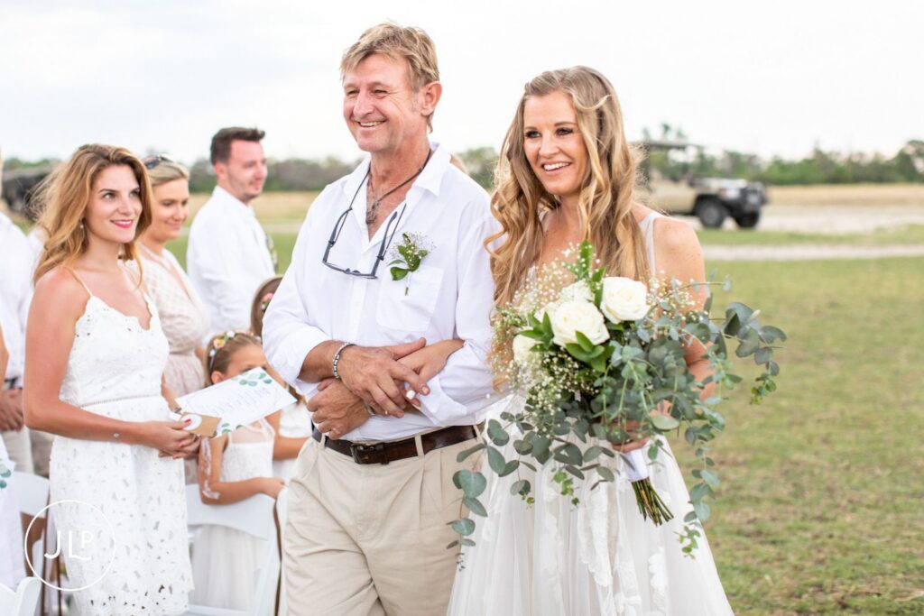 A smiling bride walks down the aisle arm-in-arm with her father at a luxury safari wedding Botswana.