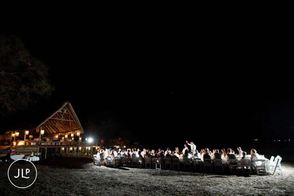 Wide night shot of guests seated at a long outdoor reception table next to an illuminated safari lodge.