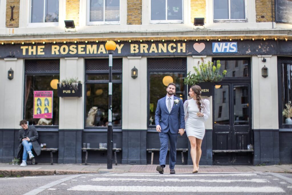 Bride and groom walking hand in hand across a zebra crossing in front of The Rosemary Branch Pub in Islington