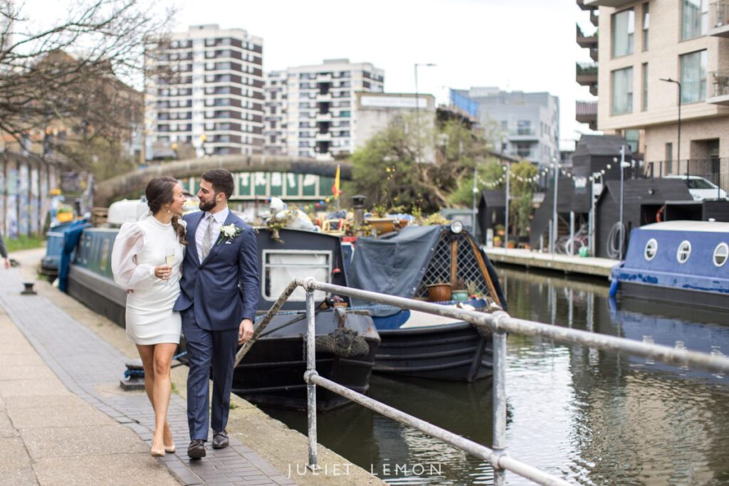 Bride and groom walking hand in hand along the bank of the canal in Islington. High rise buildings behind them and house boats on the water