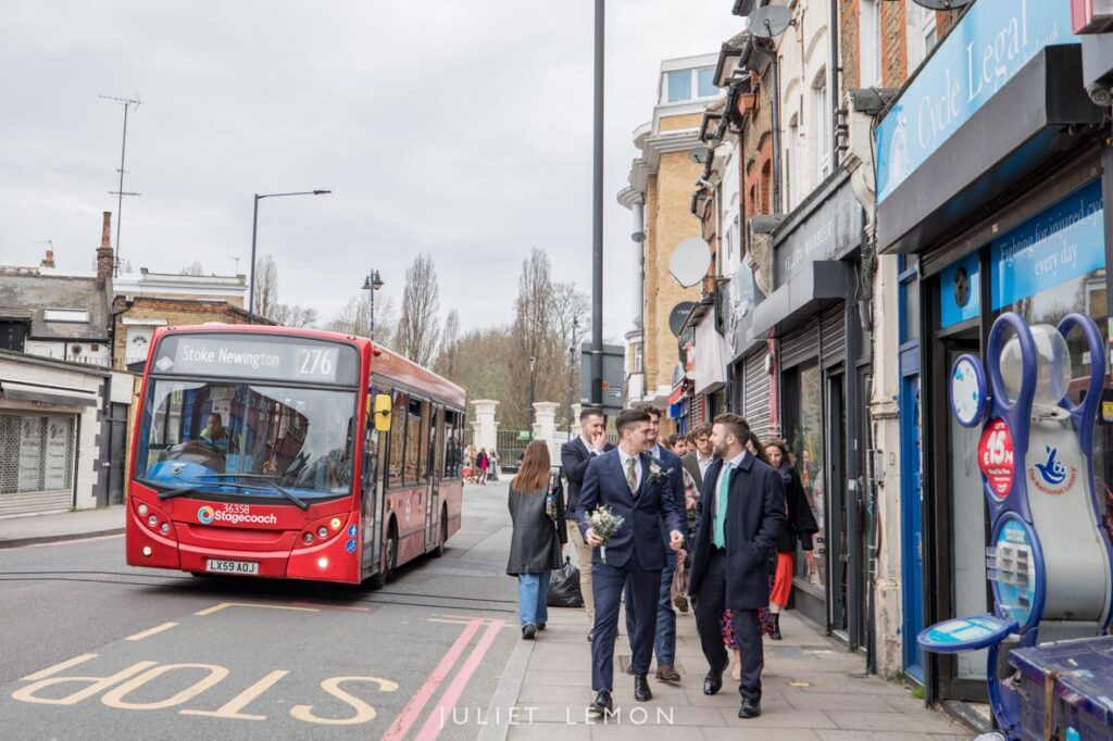 The groom and groomsmen in suits walk past shops on a Stoke Newington street
