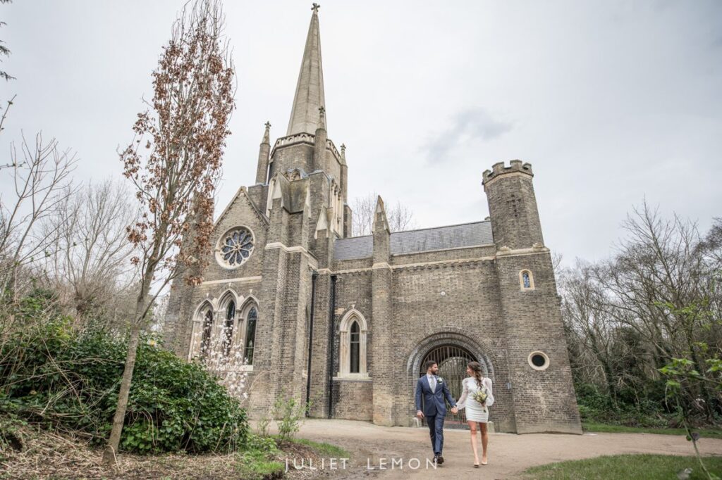 Newly married bride and groom walking hand in hand in Abney Park with Abney Park Chapel behind them in the background.
