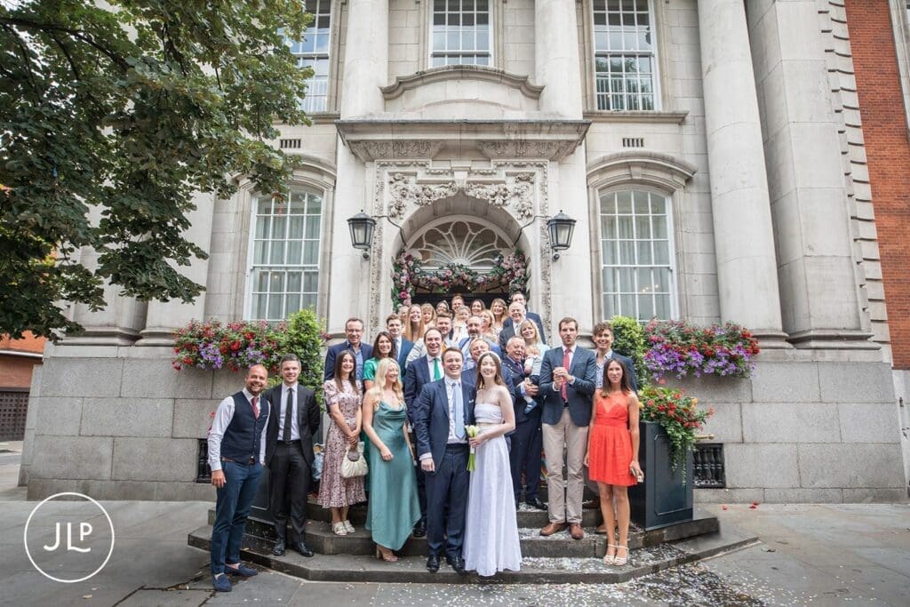 Large group wedding photo on the stone steps outside the iconic Chelsea Old Town Hall wedding venue in London.