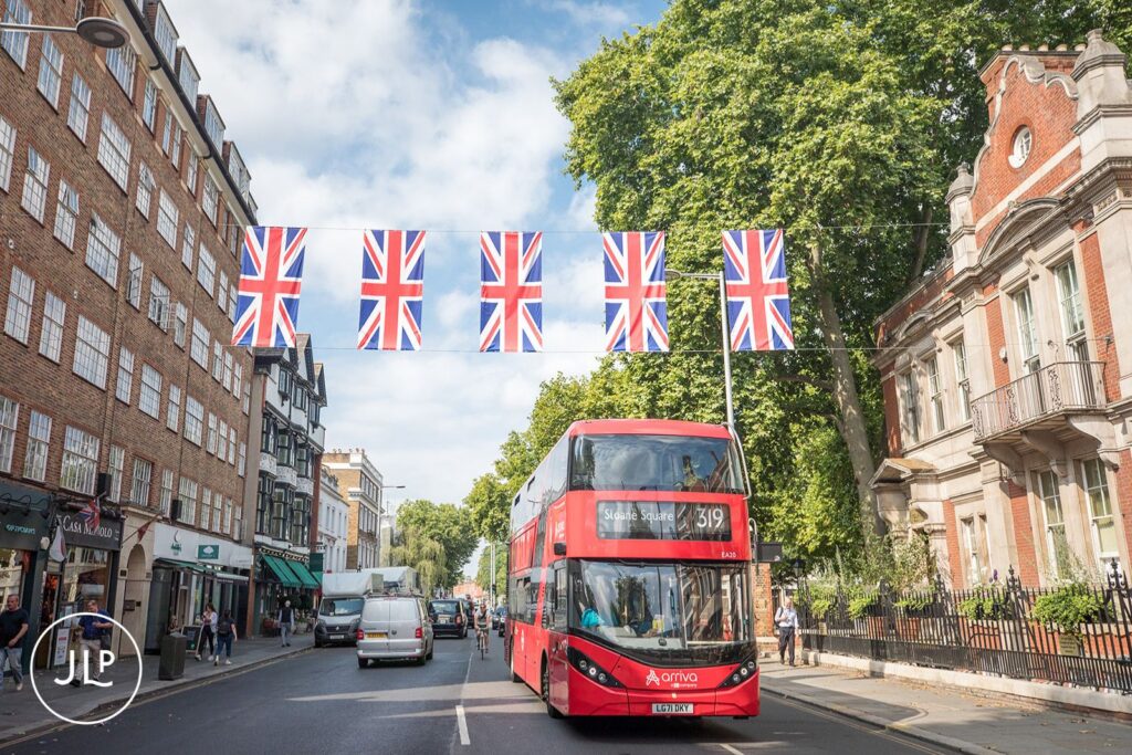 An iconic red London bus driving under Union Jack bunting near a Chelsea Town Hall wedding.