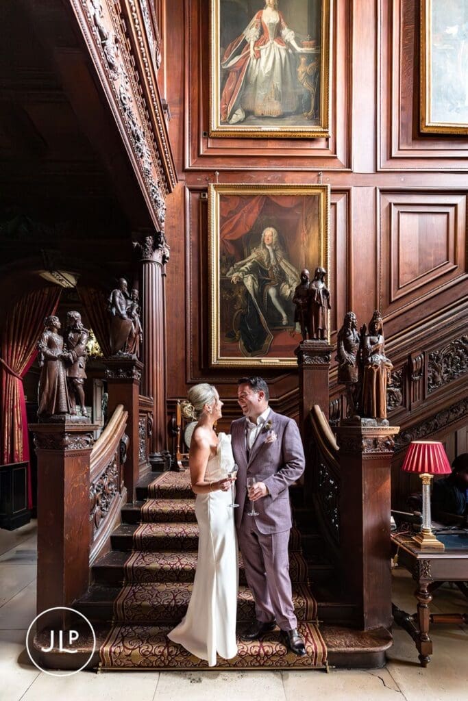 Bride and groom smiling on a grand carved wooden staircase under historic portraits during a Cliveden House wedding.