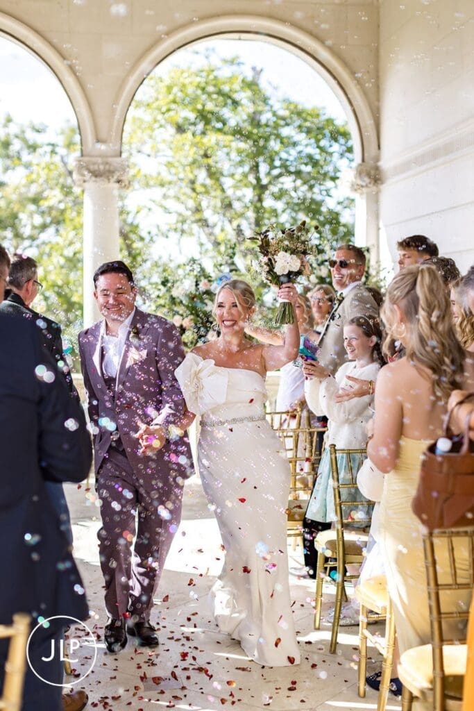 Joyful newlyweds walking through a flurry of bubbles and flower confetti, captured by a wedding photographer.