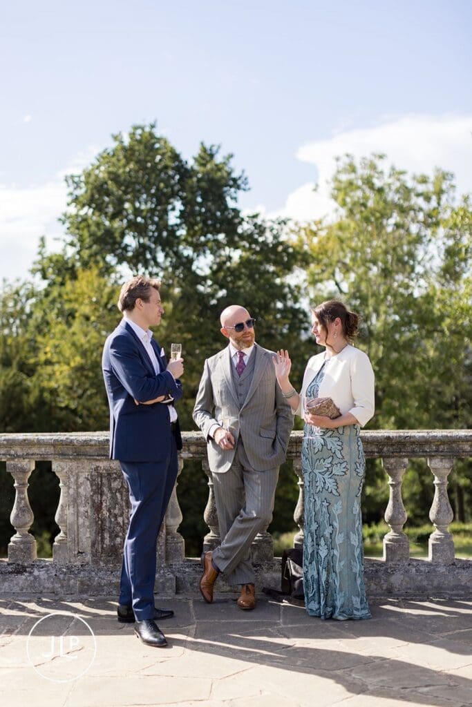 Three wedding guests chatting and holding drinks against a stone balustrade during an outdoor terrace reception.