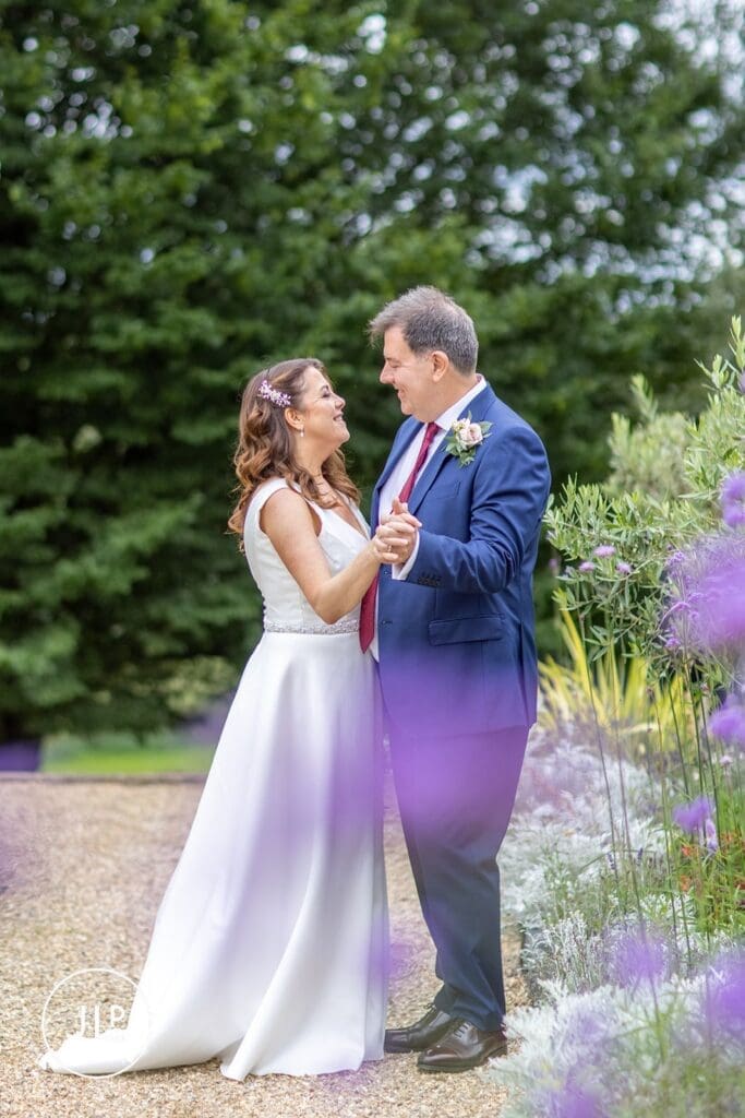 Bride and groom holding hands and smiling by purple flowers, shot by a wedding photographer in Surrey.