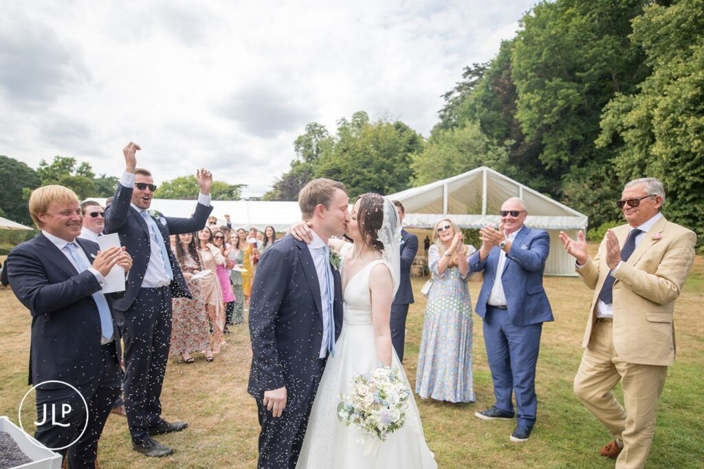 Bride and groom kissing as guests throw confetti, captured by a Surrey marquee wedding photographer.