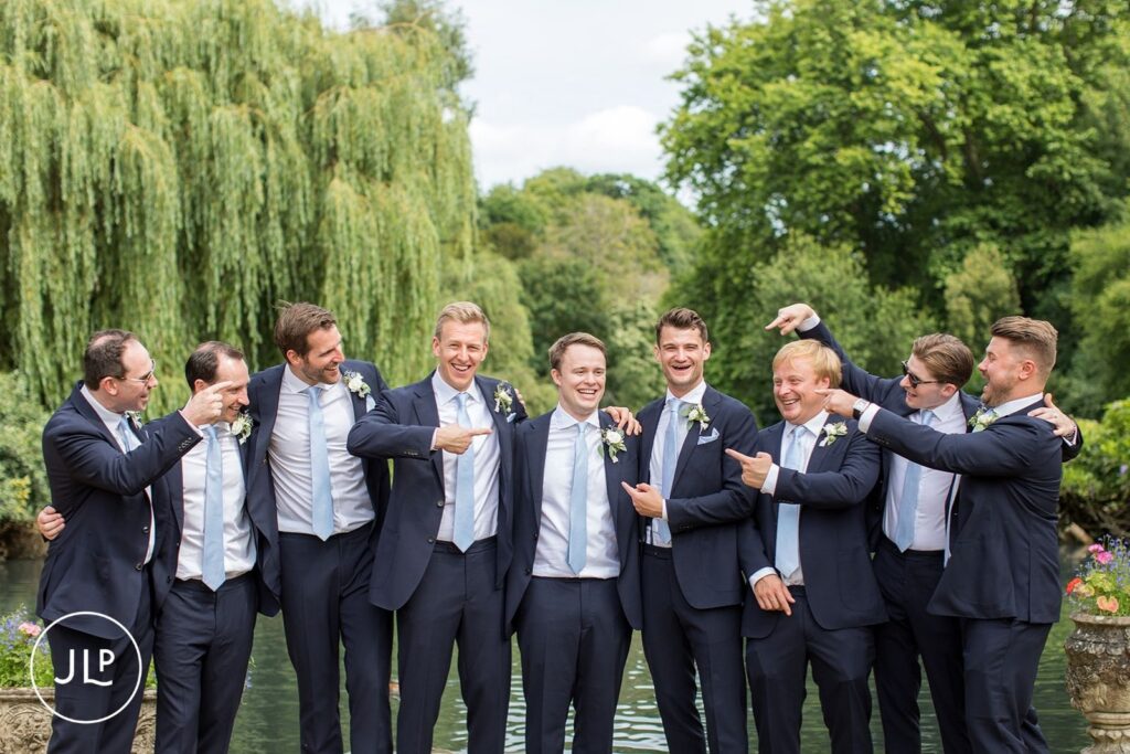 Groom and groomsmen in navy suits sharing a laugh by the lake, captured by a Surrey marquee wedding photographer.