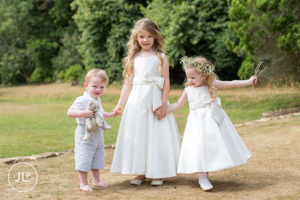Two young flower girls in white dresses holding hands with a small page boy carrying a teddy bear.