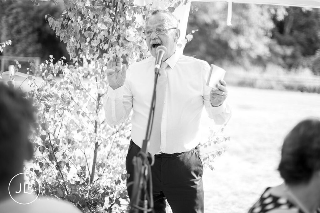 Black and white candid of an older gentleman speaking into a microphone during wedding reception toasts.