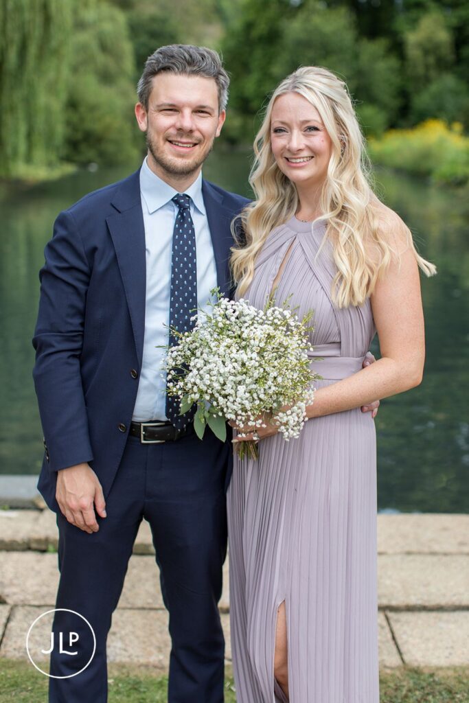 A man in a blue suit and a woman in a pleated lavender dress smiling together at a riverside wedding.