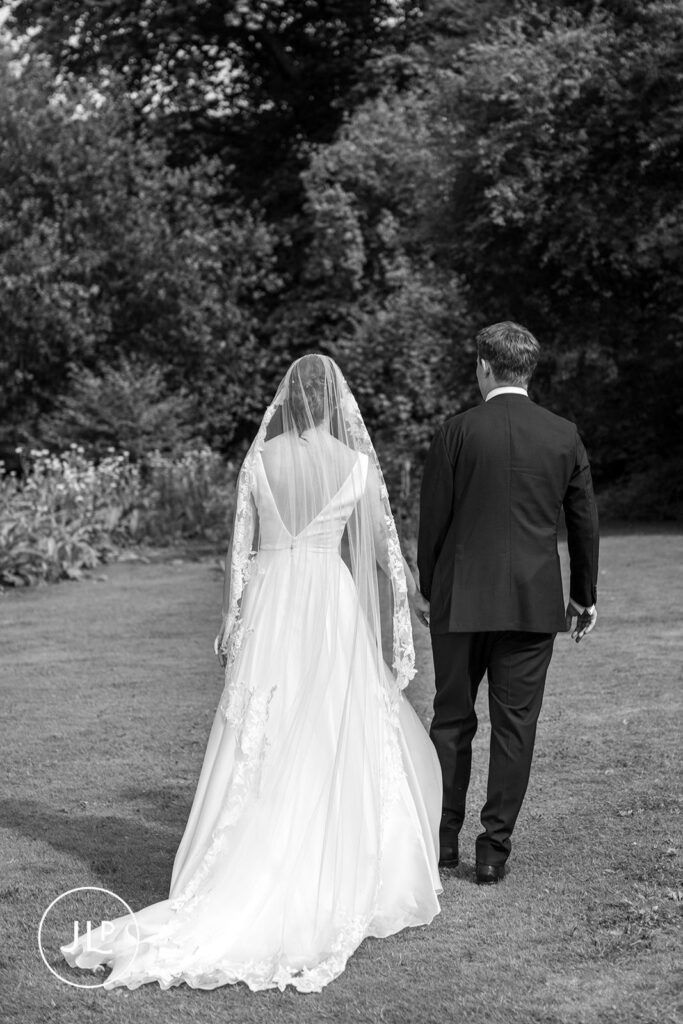 Black and white shot of the bride and groom walking away across the grass, showcasing her elegant lace veil.