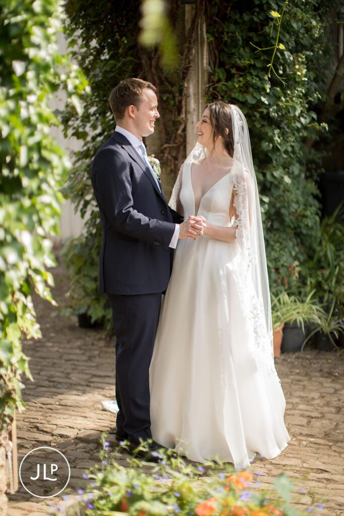 Radiant bride and groom smiling at each other in a sun-drenched courtyard during a Surrey countryside wedding.