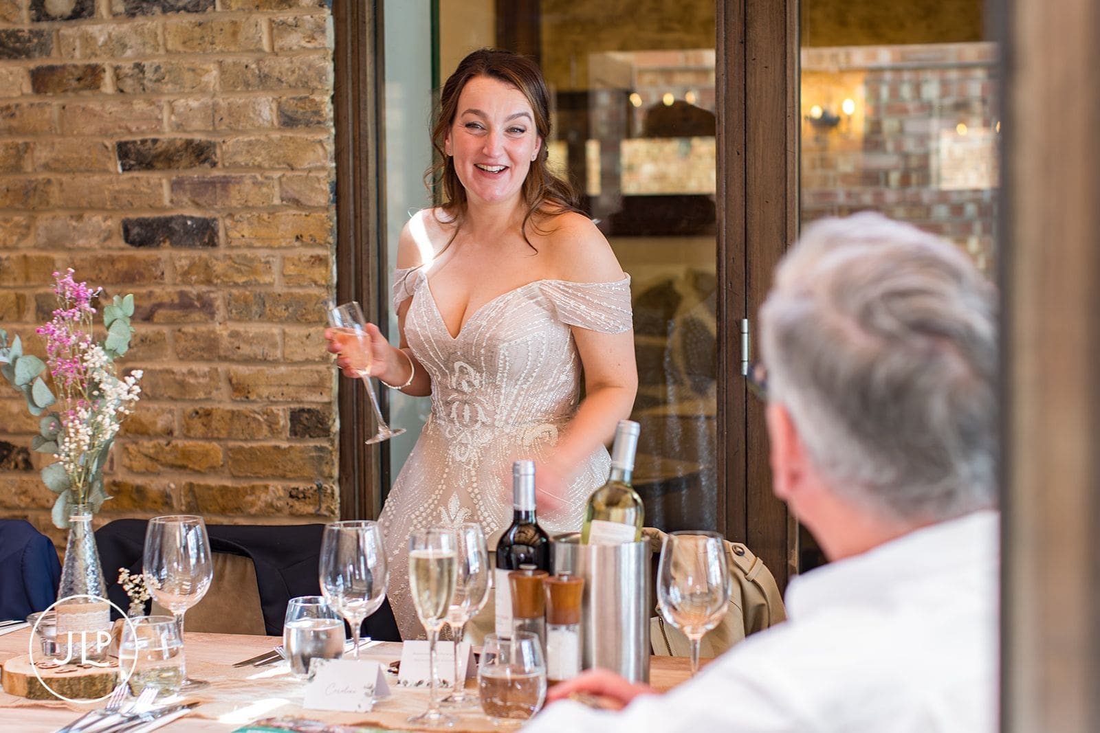 A smiling bride stands to raise a glass and make a speech during an intimate wedding breakfast.
