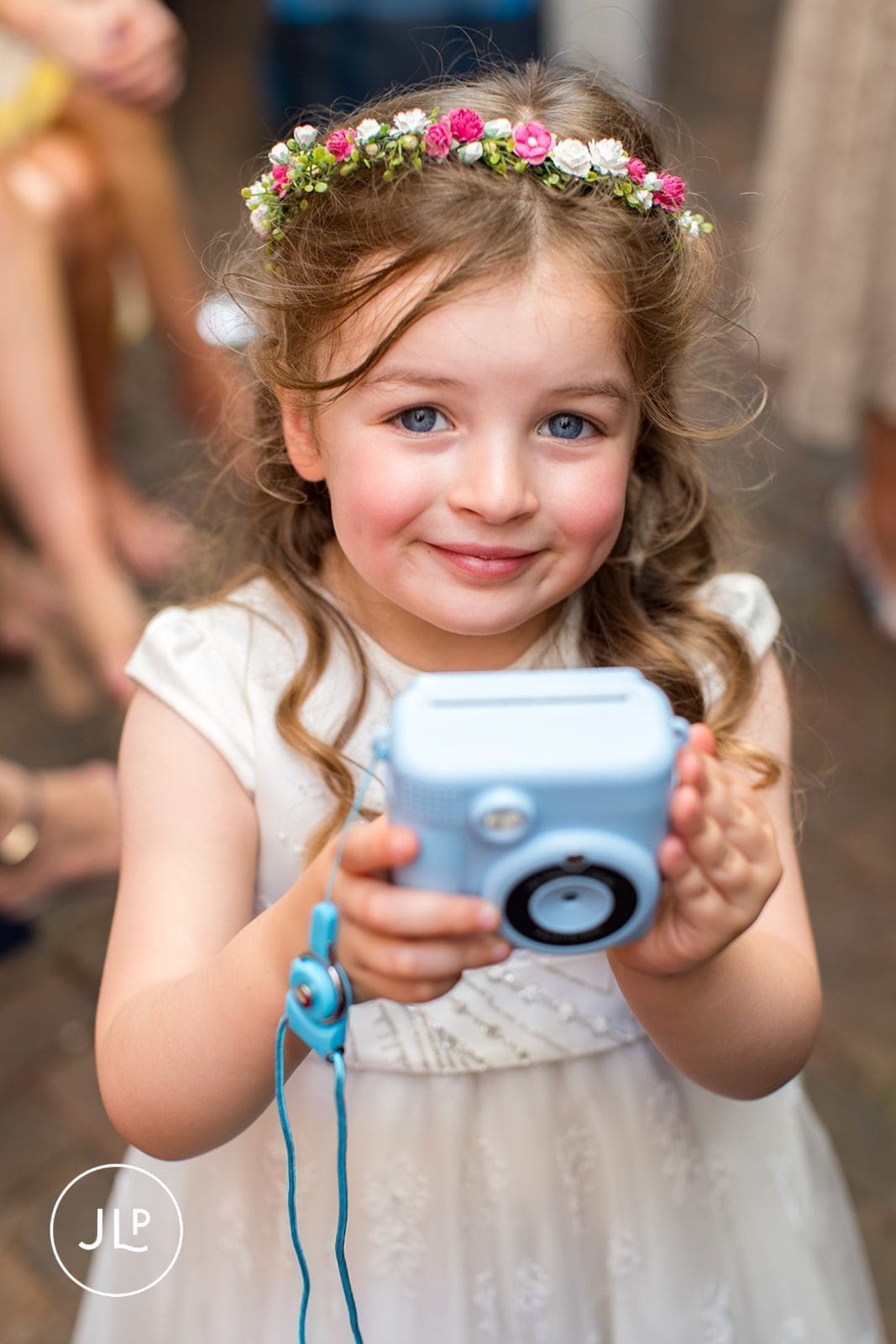A young flower girl wearing a floral crown smiles while holding up a light blue instant camera.