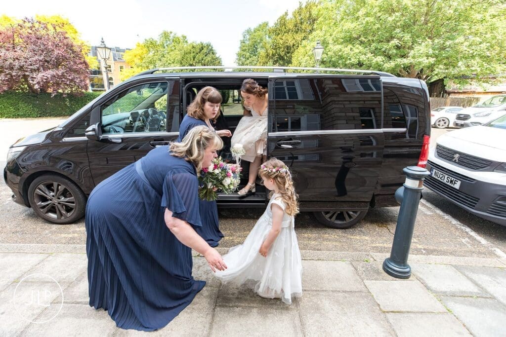 A bridesmaids in a navy blue dress helps a young flower girl adjust her skirt outside the venue before the service.