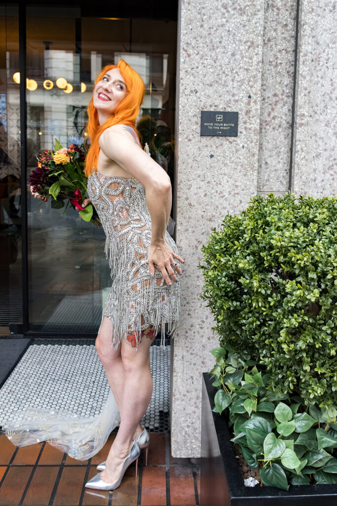 A bride with bright orange hair wearing a silver beaded fringe wedding dress poses outside a London hotel entrance.
