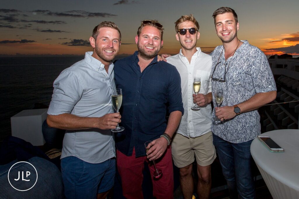 Groomsmen enjoying drinks at a pre-wedding party in Portugal with a sunset ocean view.