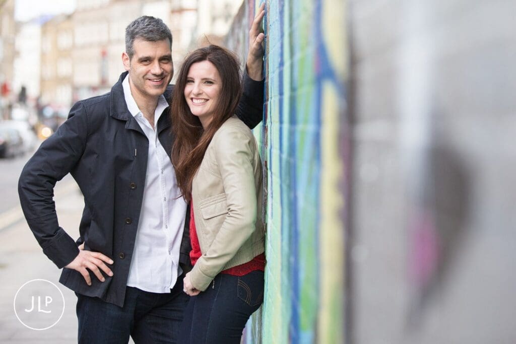 Documentary engagement photography of a couple leaning against a vibrant graffiti wall.