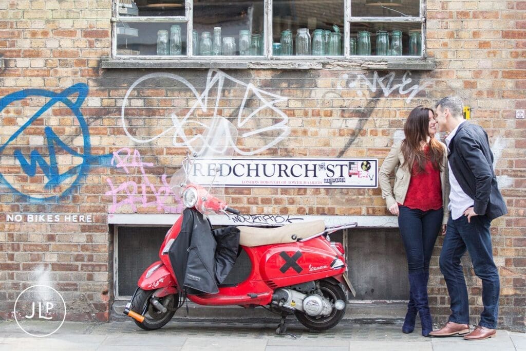 Couple leaning on a brick wall by a red Vespa, styled by a London engagement photographer.