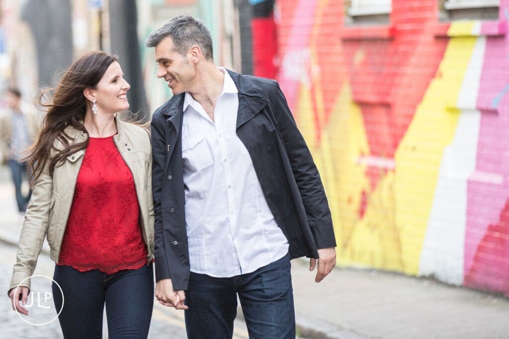 Mid-shot of a smiling couple walking past colorful murals, by a Brick Lane engagement photographer.