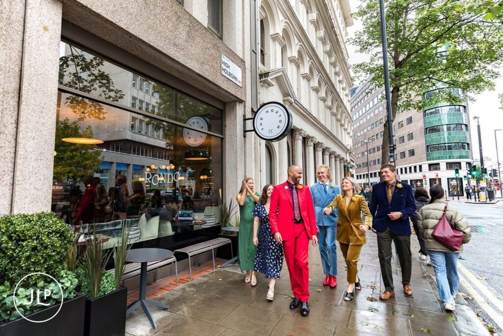 The wedding party walks together past The Hoxton hotel entrance on High Holborn street in London.