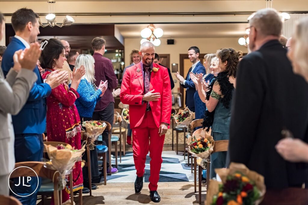 The groom in a vibrant red suit walks down the aisle during a ceremony at a London hotel.