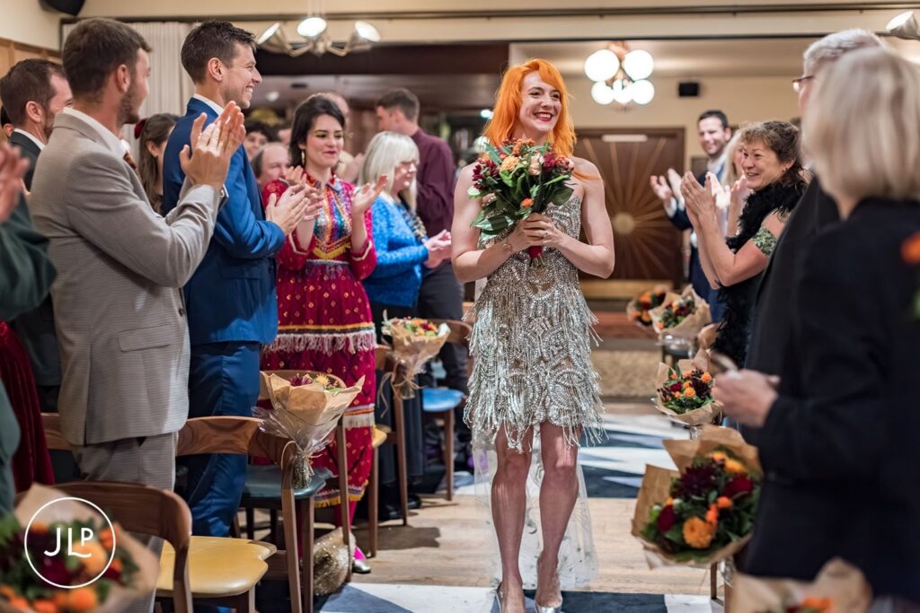 A bride with orange hair in a silver beaded dress walks down the aisle at The Hoxton Holborn.