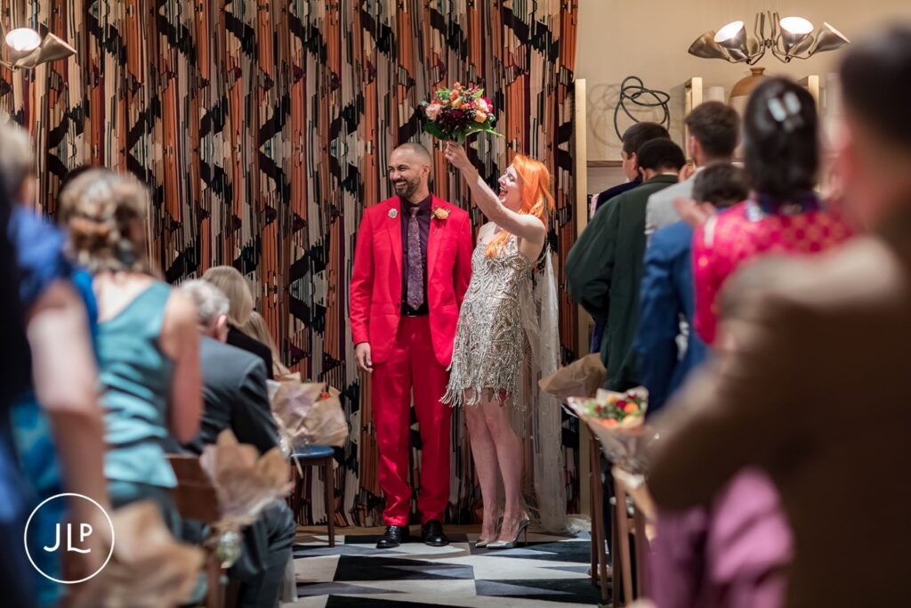 A groom in a red suit and a bride in a beaded fringe dress celebrate after saying their vows.