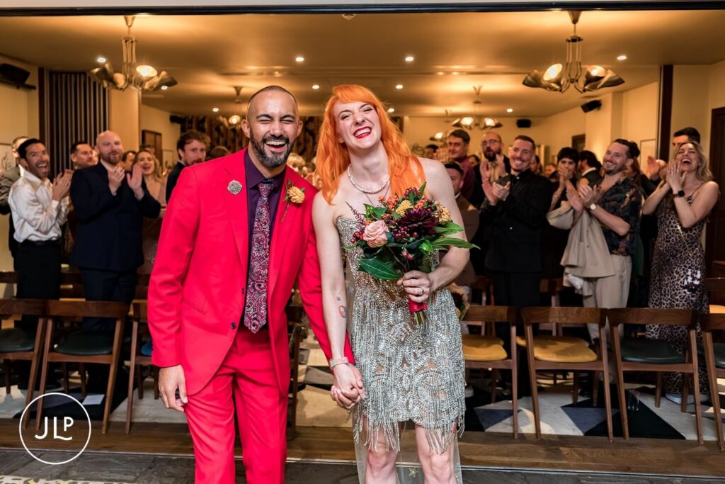 A bride with orange hair and a groom in a red suit hold hands and laugh after their wedding ceremony.