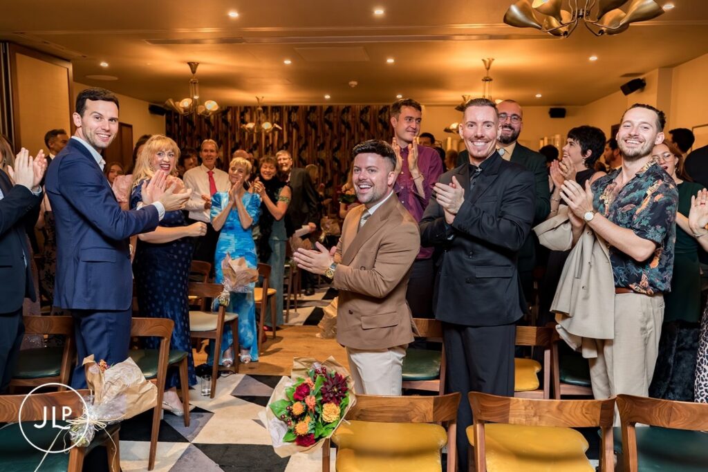 Wedding guests in stylish attire clap and cheer during a ceremony at The Hoxton Holborn in London.