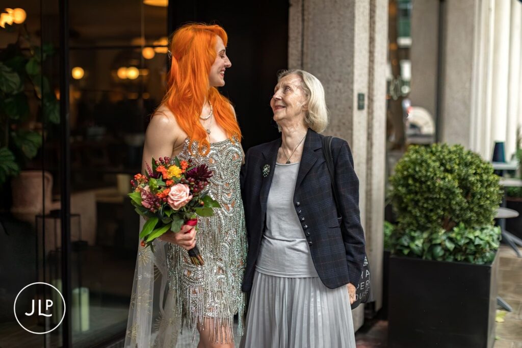 A bride with orange hair and a colorful bouquet smiles at an older wedding guest outside a London venue.