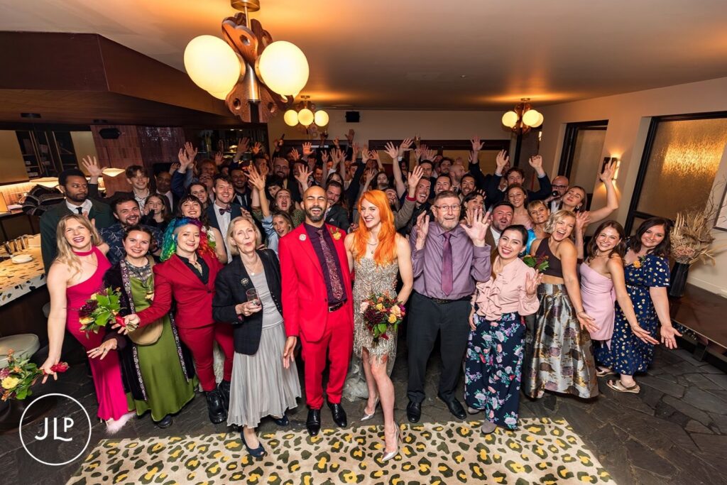 A large group of wedding guests cheers and waves while posing with the newlyweds at a London hotel bar.