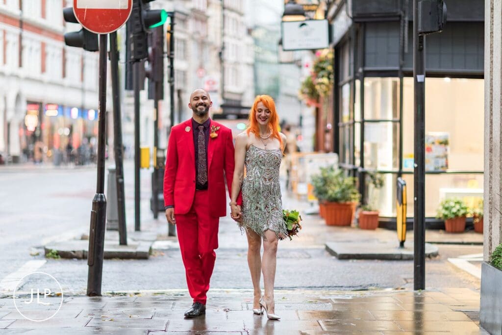 The bride and groom walk hand-in-hand past a red traffic light on a rainy afternoon in Holborn, London.