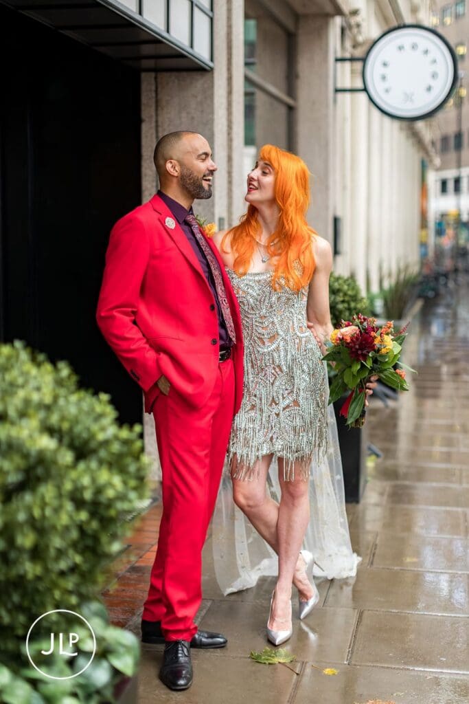 A groom in a vibrant red suit and a bride with orange hair laugh together outside a stylish London hotel.