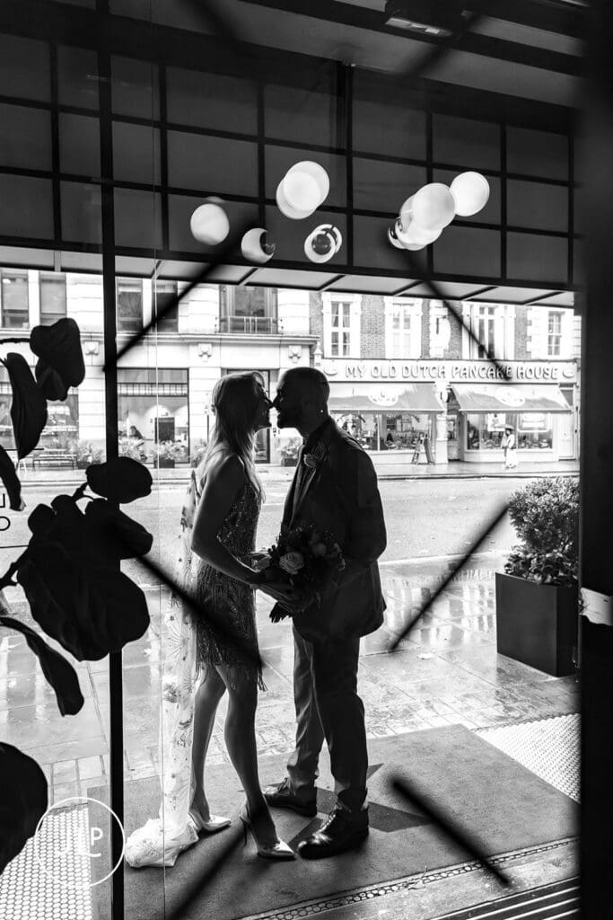 A black and white silhouette of a bride and groom kissing inside the glass doors of a London hotel.