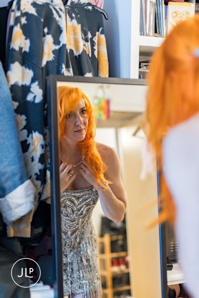 A bride with vibrant orange hair checks her reflection in a mirror while getting ready in Putney.