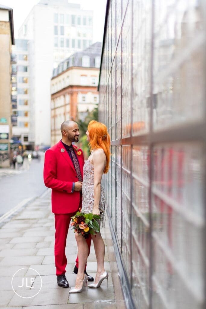 A bride in a short sparkly dress and a groom in a red suit look at each other on a rainy London street.