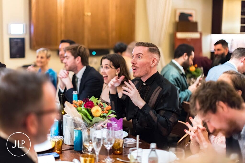 A guest with short hair and a black mesh shirt talking animatedly at a wedding reception table adorned with colorful flowers.